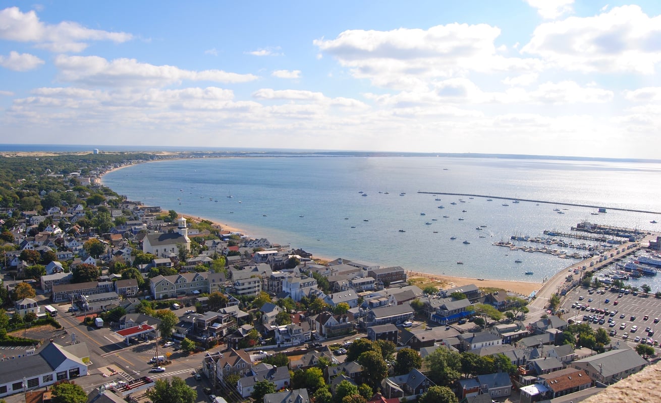 Aerial view of a coastal town with buildings, a curved shoreline, a harbor with boats, and a blue sky with scattered clouds—an inviting scene that highlights why such places are sought-after travel destinations.