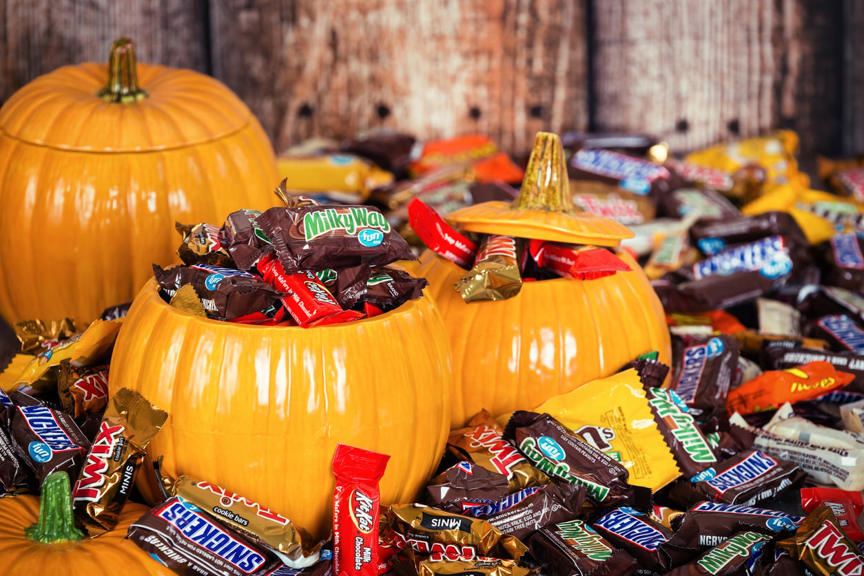 Ceramic pumpkins filled with assorted Halloween candy bars, such as Milky Way, Snickers, and KitKat, sit surrounded by more wrapped candies on a rustic wooden surface.