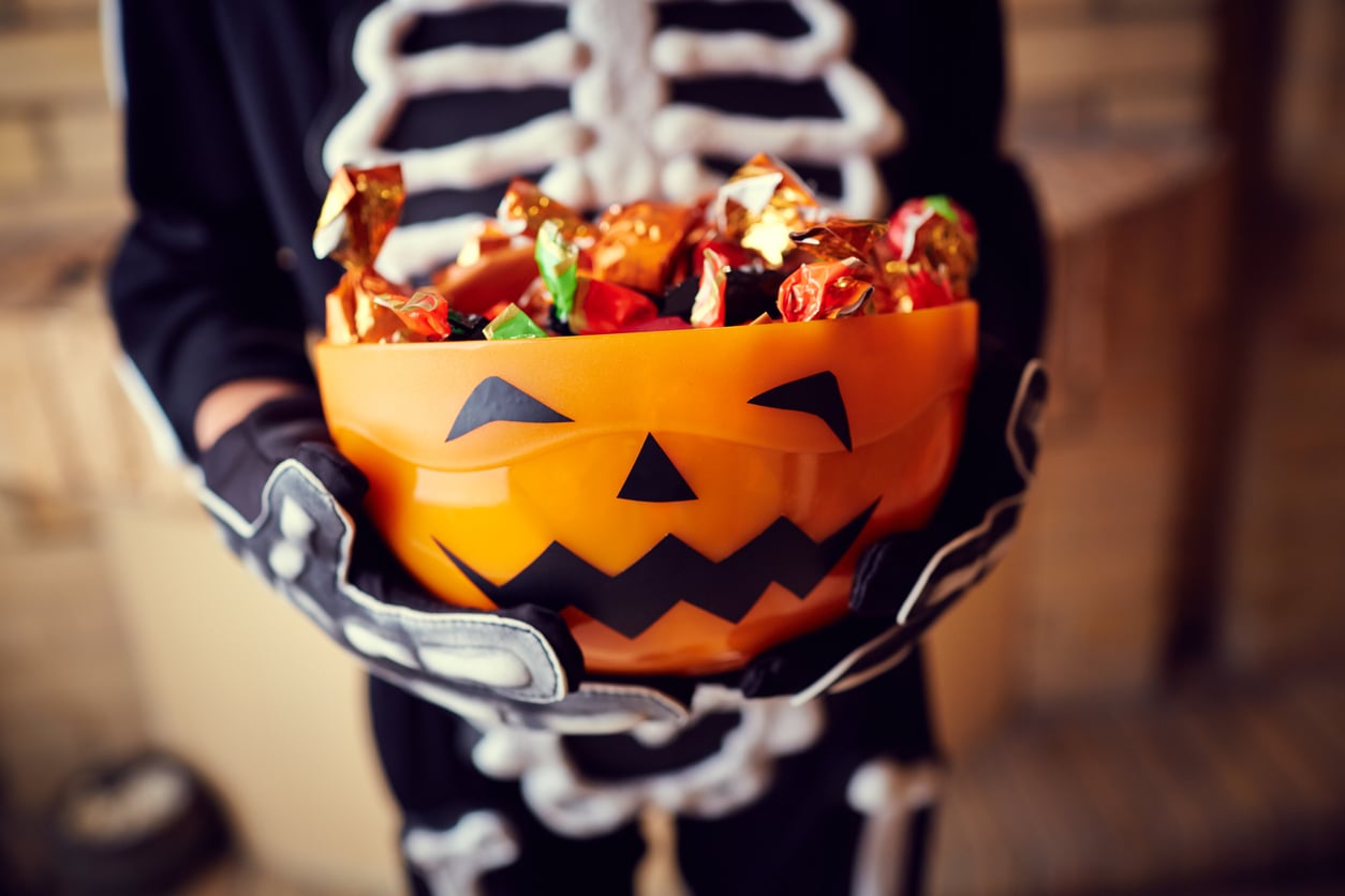A person in a skeleton costume holds a pumpkin-shaped bucket filled with wrapped Halloween candy.
