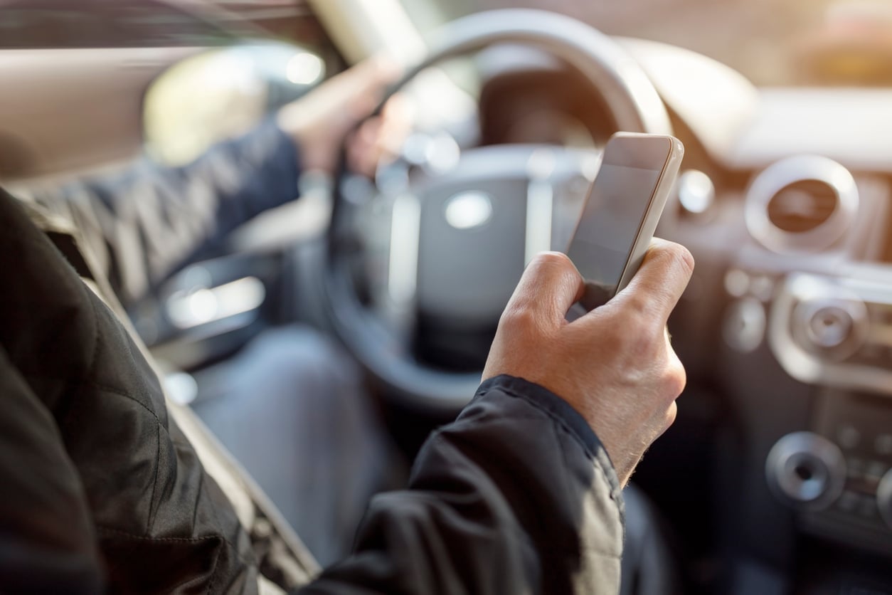 A person driving a car holds the steering wheel with one hand and uses a smartphone with the other hand, suggesting distracted driving.