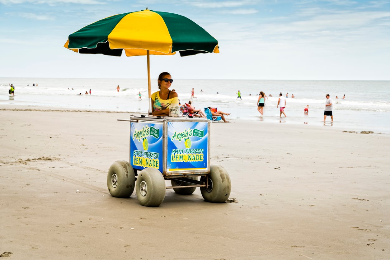 A woman sits under a green and yellow umbrella on a lemonade cart with large wheels at the beach, enjoying her beach vacation savings. People are swimming and walking in the background near the ocean.