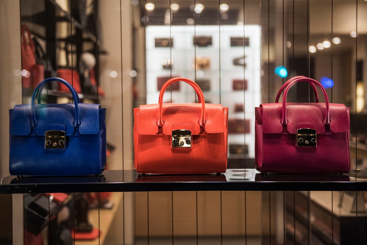 Three handbags—blue, orange, and magenta—are displayed side by side on a black shelf in a brightly lit store, with shelves of other bags blurred in the background, offering a glimpse into products people no longer buy as frequently.