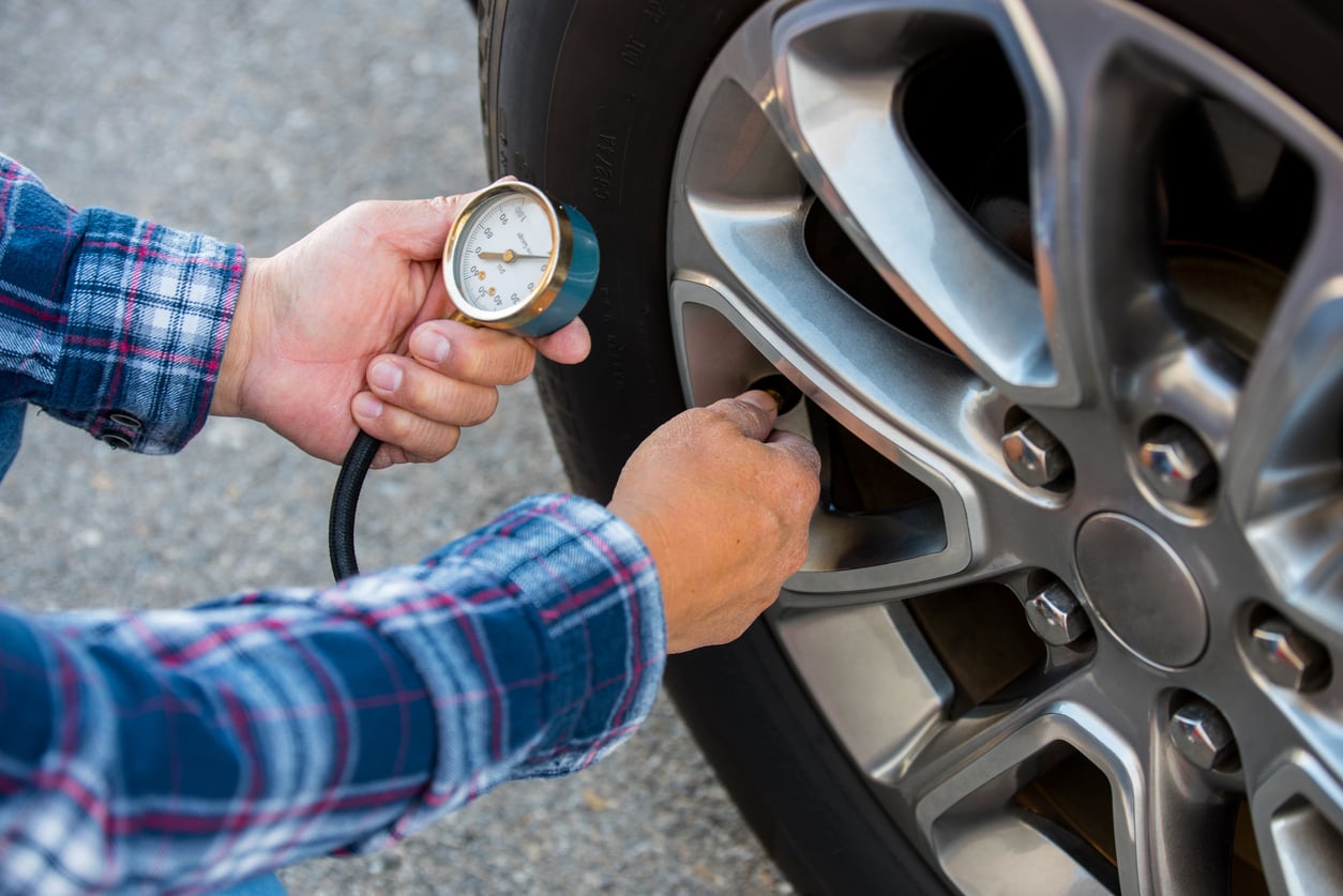 A person in a plaid shirt checks a car tire’s air pressure using a tire pressure gauge, holding the gauge to the valve on the wheel—a crucial step in any road trip checklist.