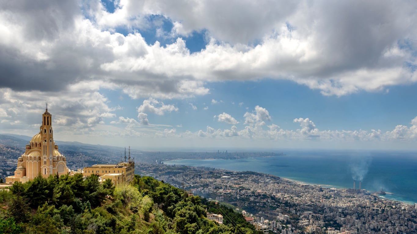A church on a hill overlooks a coastal city with a blue sea, distant cityscape, clouds in the sky, and smoke rising from part of the shoreline—creating a picturesque scene that makes it one of the region’s must-see travel destinations.