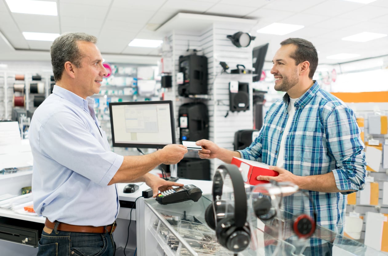 A man at a store counter pays with a credit card while the cashier smiles and processes the transaction. Electronic products and accessories, including some waste of money products, are visible around them.