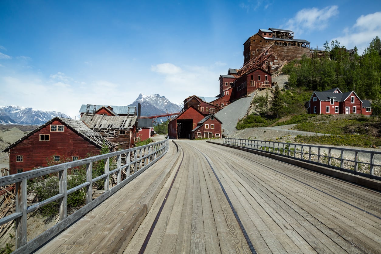 A wide wooden bridge leads to a cluster of old red buildings on a hillside, evoking the charm of historic American towns, with snowy mountains and a blue sky in the background.