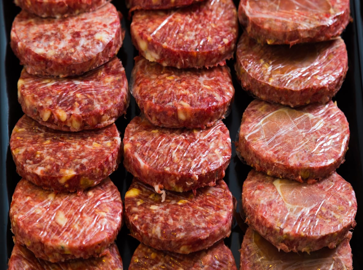 Rows of raw beef burger patties wrapped in plastic sit on a black tray, ready for cooking or grilling.