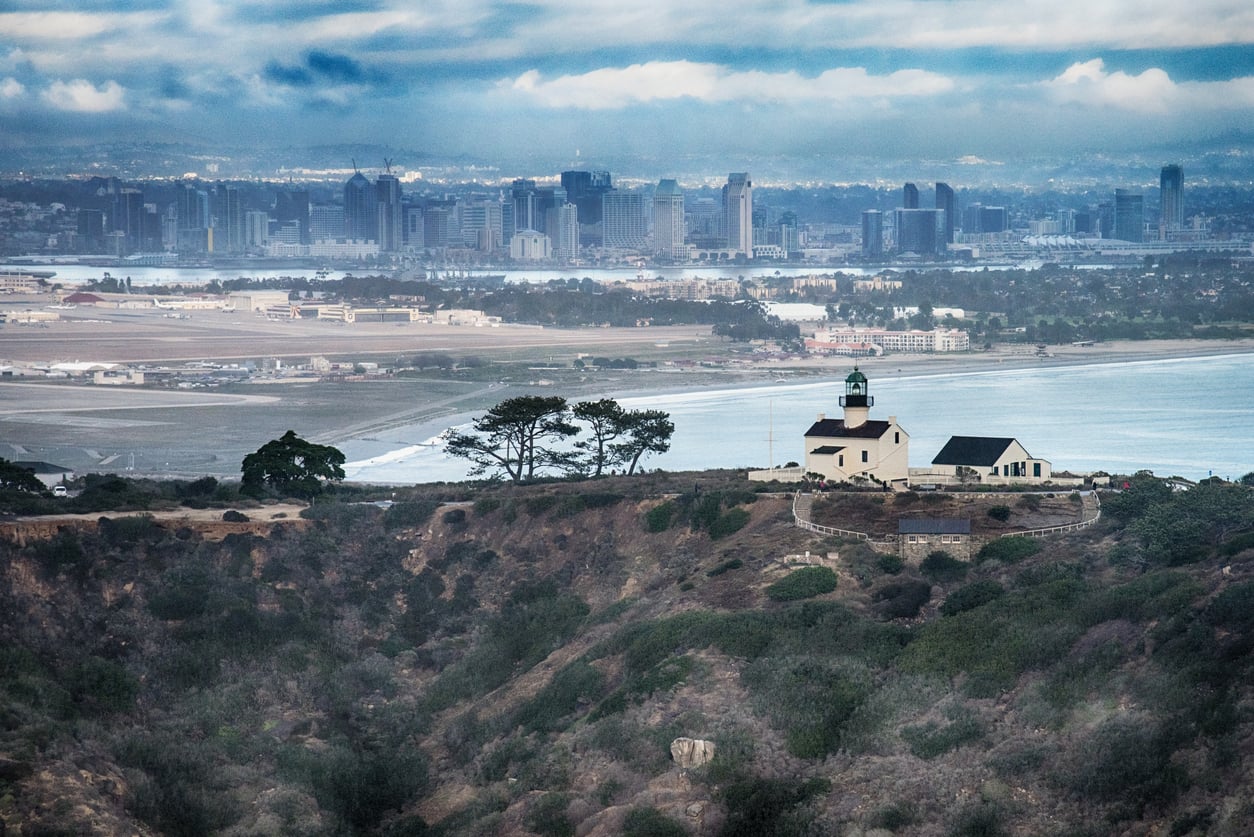 A white lighthouse sits on a hill surrounded by trees, overlooking a city skyline with tall buildings and an airport near the coast under a cloudy sky—an iconic scene among travel destinations on the West Coast.