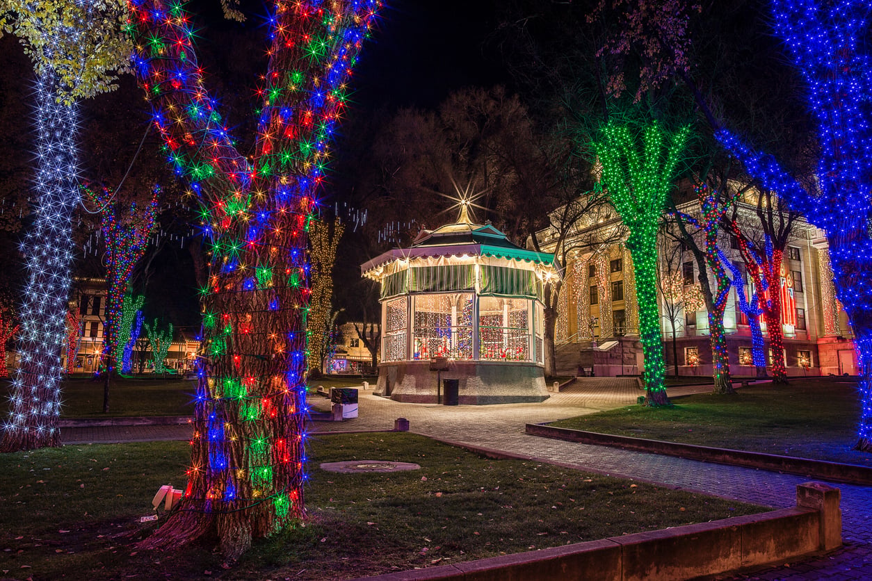 Colorful holiday lights wrap around trees and a gazebo in a park at night, with a historic building illuminated in the background. Capturing the spirit found in the best Christmas towns in America, the scene is festive and vibrant, with a mix of red, blue, green, and white lights.