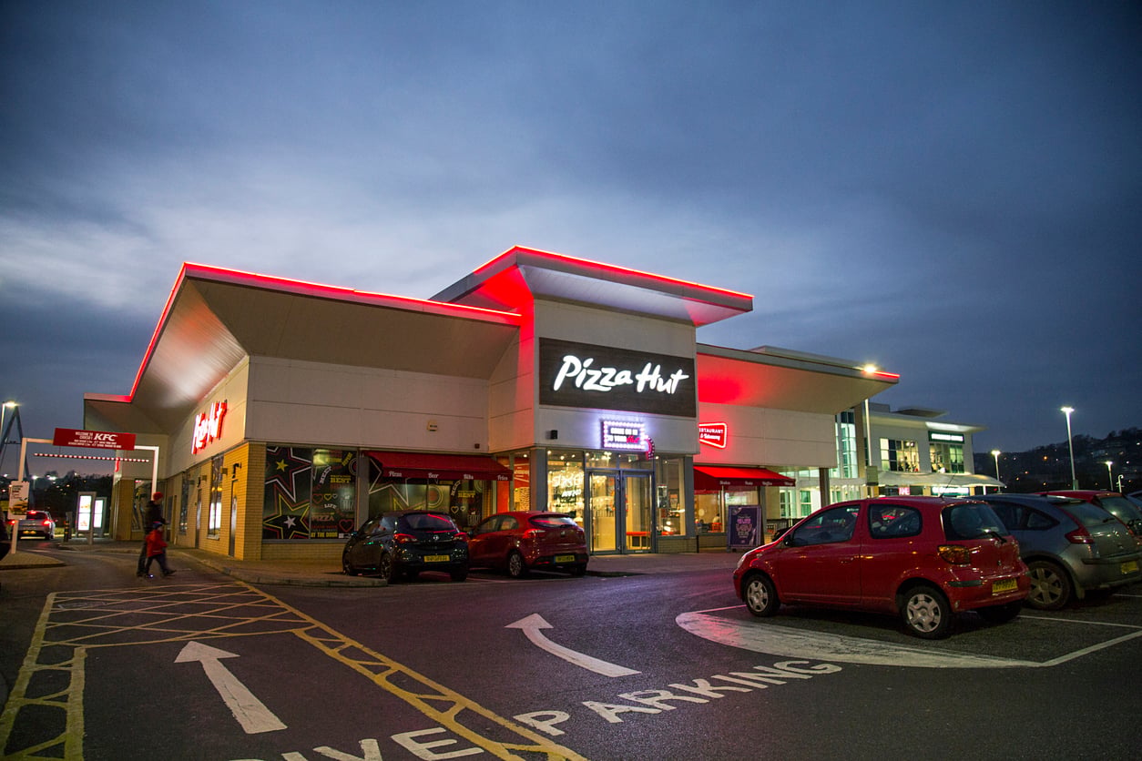 A Pizza Hut restaurant with illuminated signage at dusk, surrounded by parked cars in a parking lot. Other storefronts and streetlights are visible in the background under a cloudy sky.