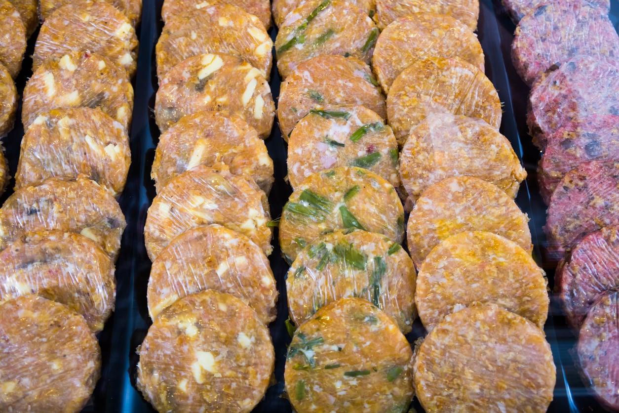 Rows of uncooked burger patties wrapped in plastic, arranged neatly in a display case. Some patties have visible chunks of vegetables or herbs mixed in.