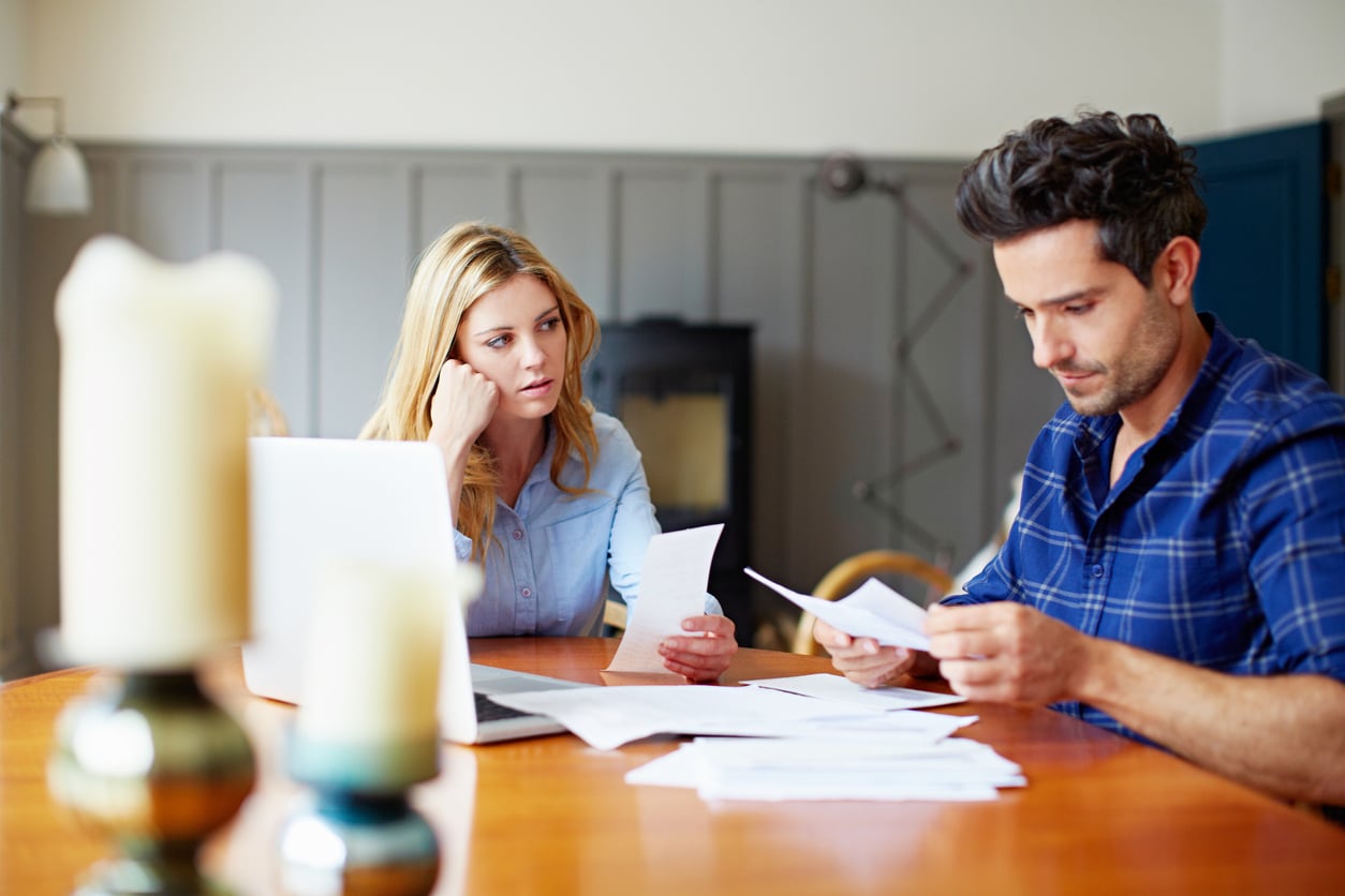 A concerned woman and a man sit at a table with papers and a laptop, reviewing documents together in a home setting.