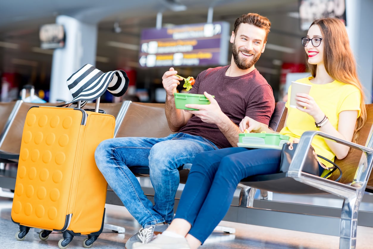 A smiling man and woman sit in an airport terminal, enjoying food from containers as they discuss their upcoming vacation swaps. A large yellow suitcase with a hat on top is beside them, while blurred departure signs appear in the background.