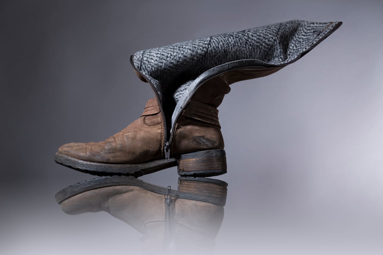 A single brown leather boot with a zipper is open, showing its gray patterned inner lining. Unlike products that don't last, this boot stands upright on a reflective surface against a plain background, highlighting its quality construction.