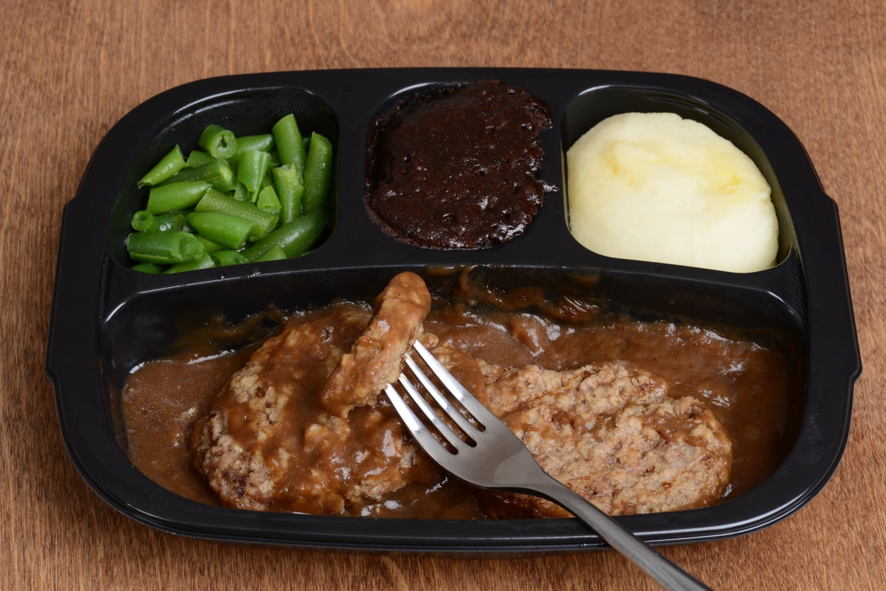 A TV dinner with Salisbury steak in brown gravy, green beans, mashed potatoes, and a chocolate brownie in a black plastic tray, with a fork holding a piece of steak.