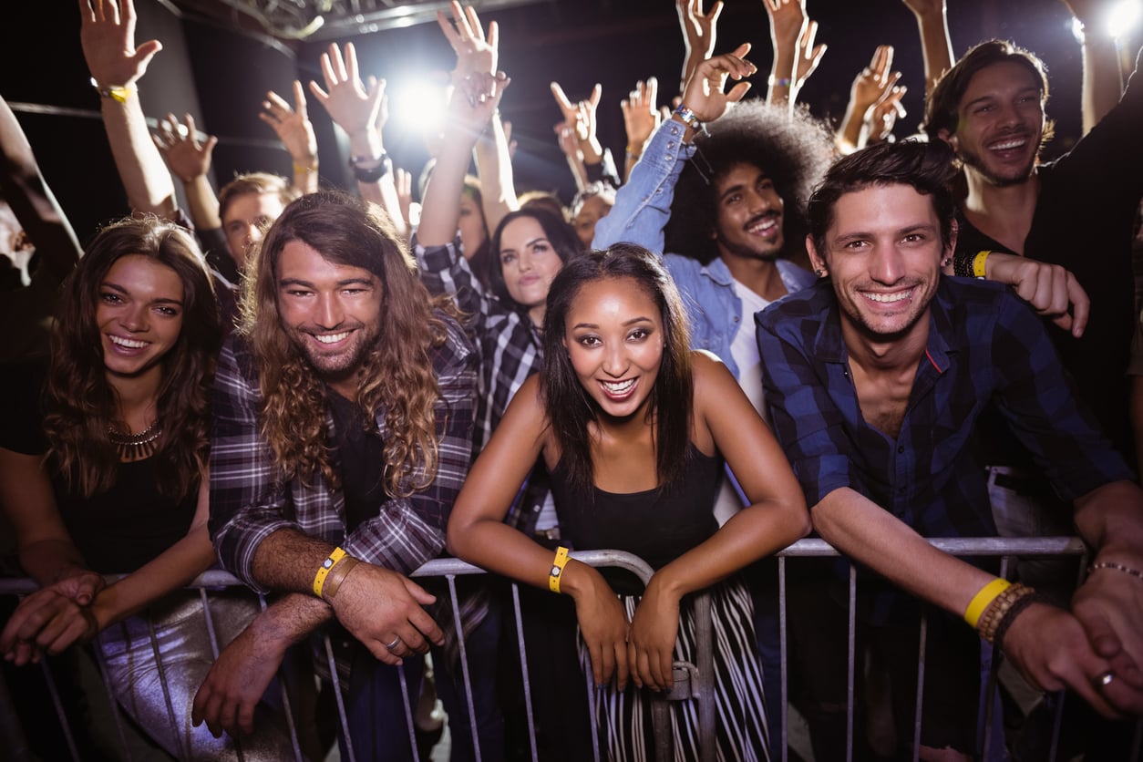 A group of young people stand at a concert barrier, smiling and raising their hands, enjoying live music at a crowded indoor event.