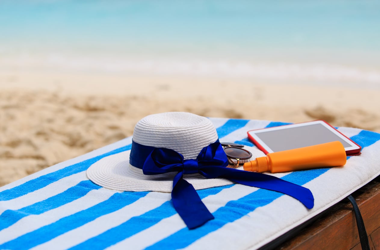 A white sun hat with a blue ribbon, sunglasses, sunscreen, and a tablet rest on a blue and white striped towel at the beach, capturing the perfect scene for beach vacation savings with sand and ocean in the background.