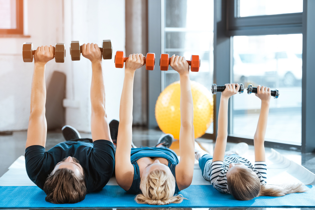 Three people—a man, a woman, and a child—lie on exercise mats indoors, raising dumbbells above their chests. Exercising together at home lets them save money while staying fit; a yellow exercise ball sits in the background.