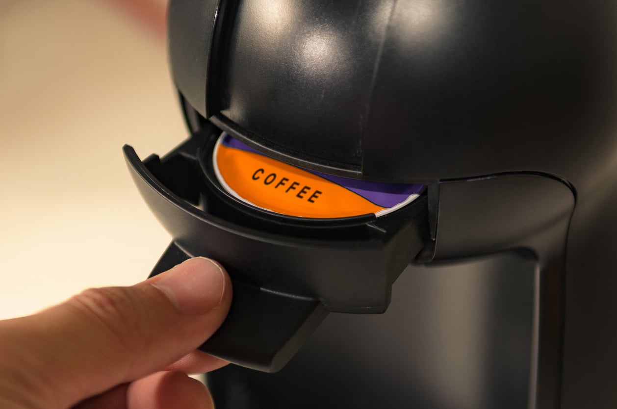 A hand inserts an orange coffee pod labeled "COFFEE" into a black single-serve coffee machine—a glimpse of products people no longer buy as tastes and trends evolve.