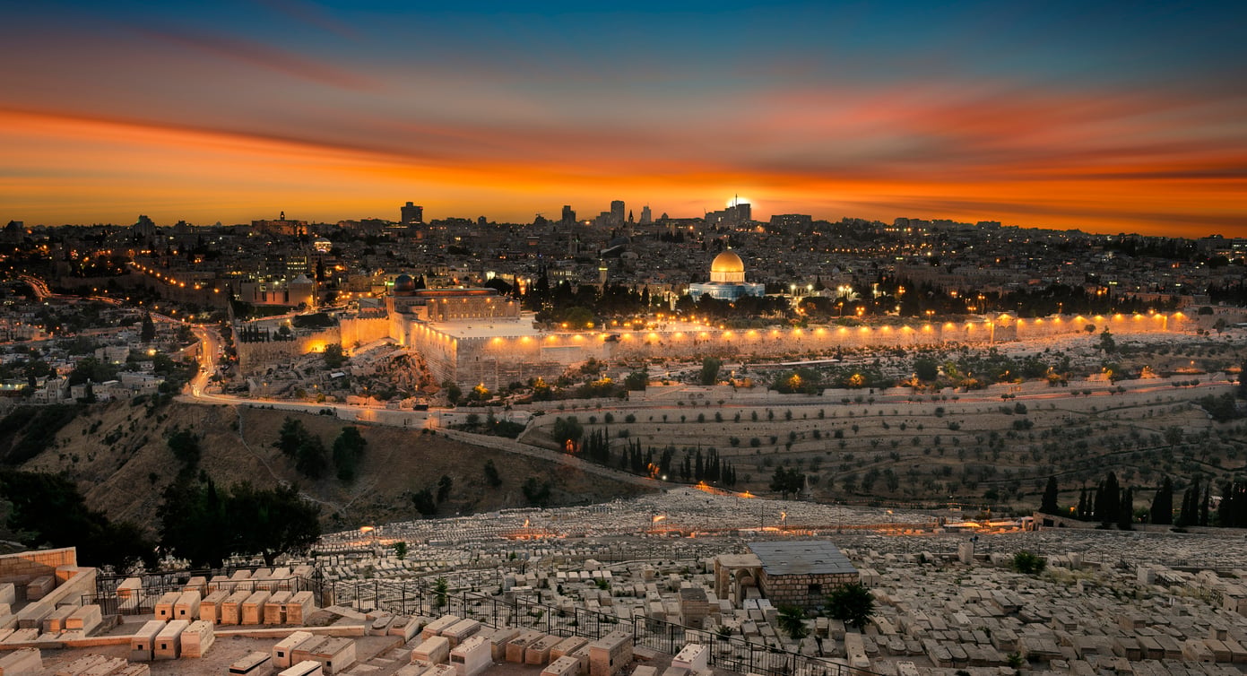 A panoramic view of Jerusalem at sunset, featuring the Dome of the Rock, city lights, and the Mount of Olives cemetery in the foreground under a colorful sky—an unforgettable scene that highlights why Jerusalem remains one of the world’s most captivating travel destinations.