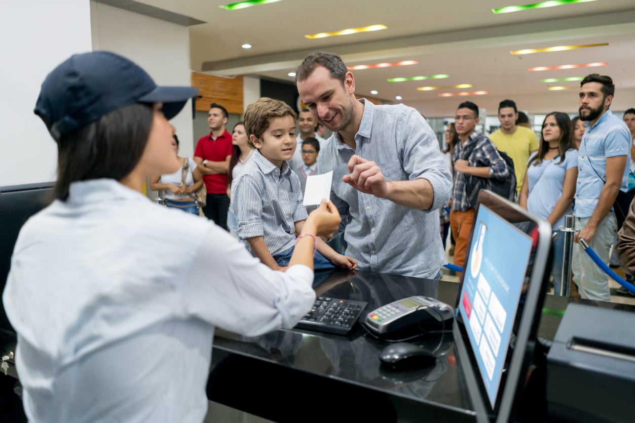 A man and a young boy smile as they receive tickets from a female cashier at a counter, using movie theater hacks, with a long line of people waiting behind them in a brightly lit indoor setting.