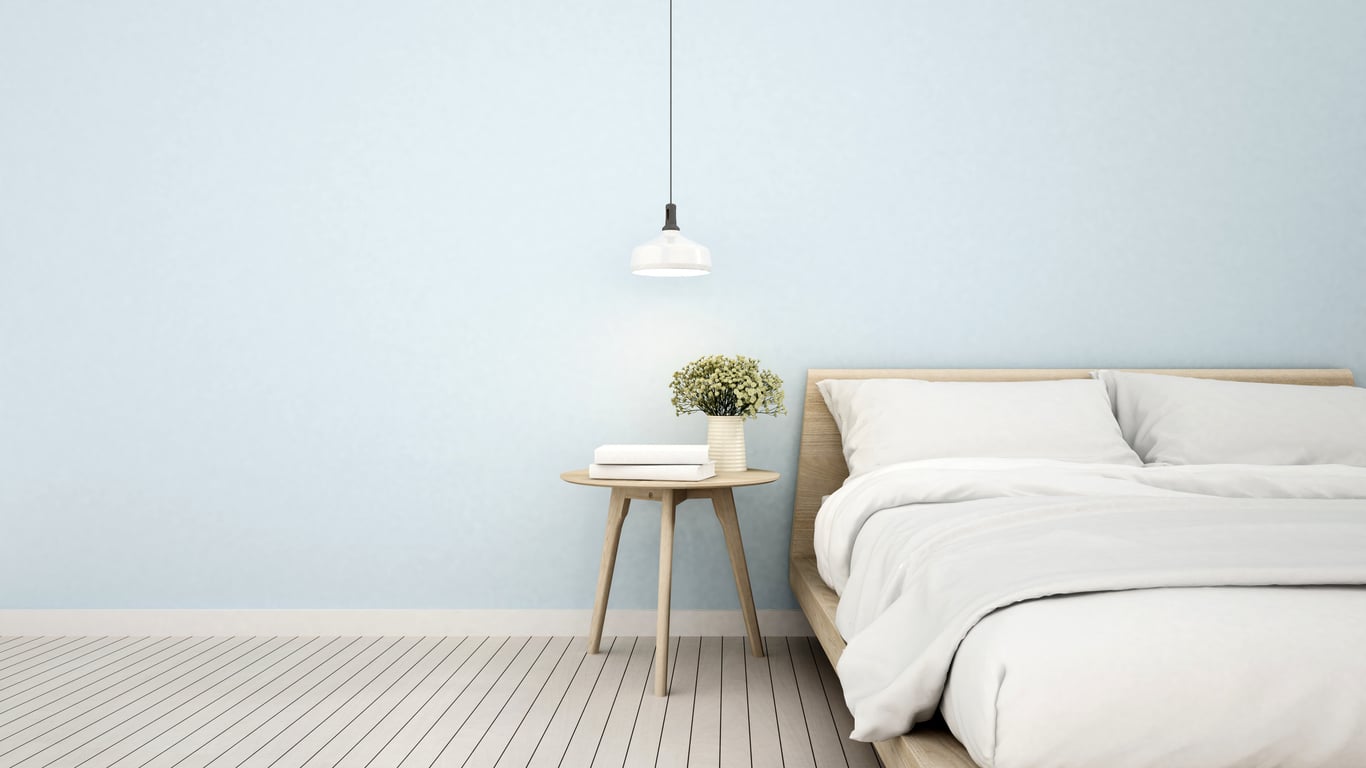 Minimalist bedroom with light blue walls, a wooden bed with white bedding, and a small round bedside table holding books and a potted plant—ideal for embracing hotel changes in décor trends. A white pendant light hangs overhead.