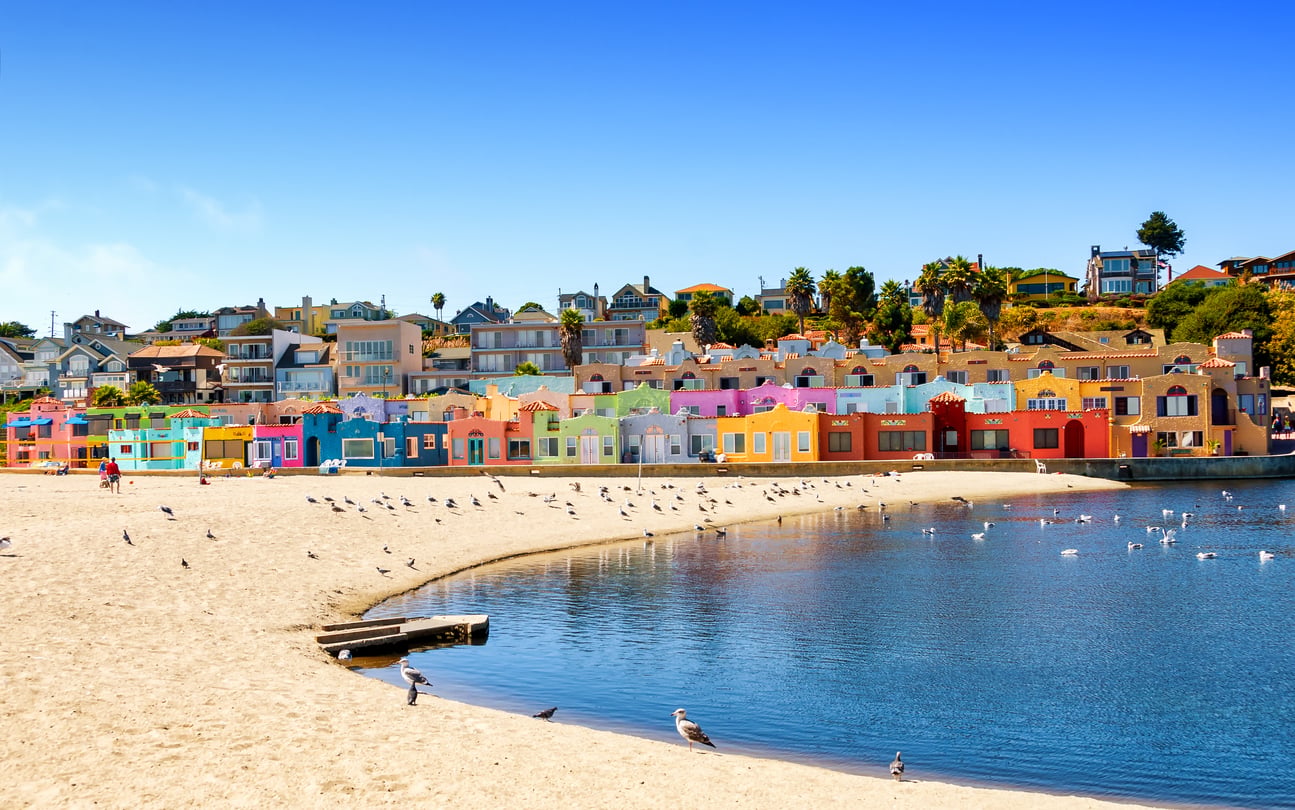 Colorful houses line the sandy shore of a calm beach, capturing the charm often found in picturesque beach towns, with seagulls on the sand and in the water, under a clear blue sky.