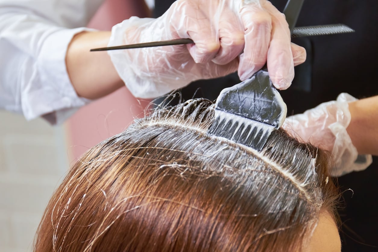 A person applies hair dye to the roots of someone’s brown hair using a brush; both hands wear clear gloves, and a comb is visible in the background.