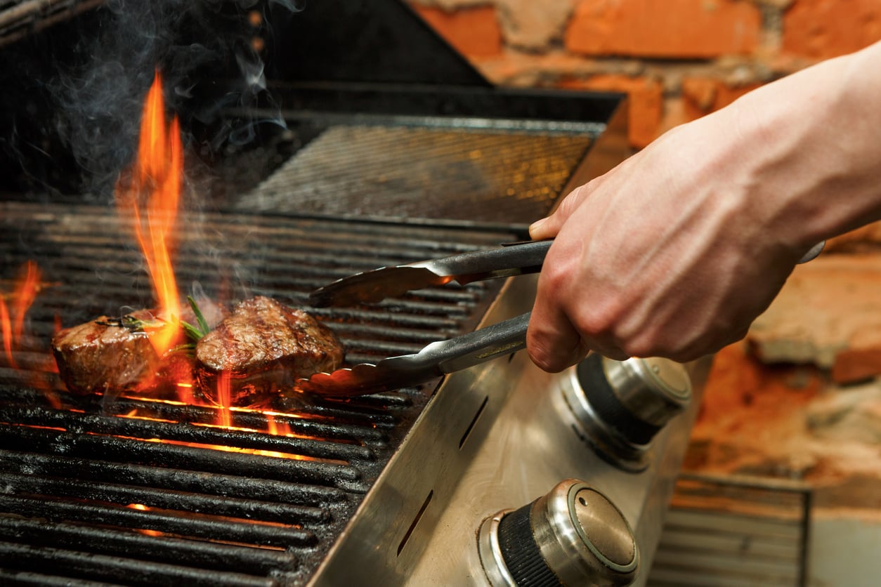 A person uses tongs to grill two steaks over an open flame on a barbecue grill, with a brick wall in the background.
