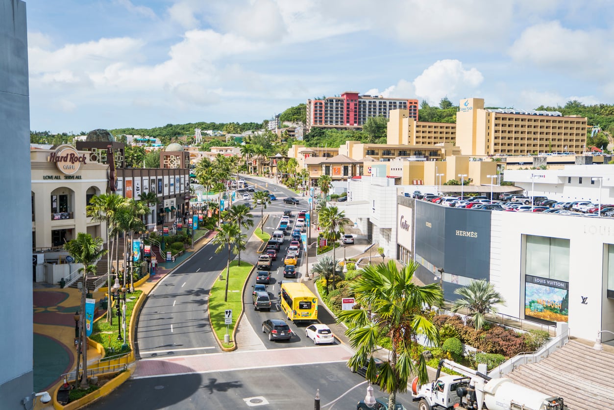 A sunny day in a bustling city with busy streets, palm trees, cars in traffic, and buildings including shops, hotels, and a Hard Rock Café—one of the iconic travel destinations for visitors. A hill with more buildings is visible in the background.