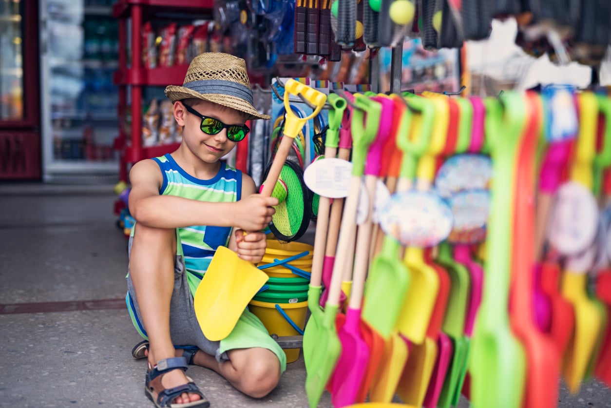 A young boy wearing a hat, sunglasses, and a striped tank top sits on the ground in a store, smiling and holding a yellow plastic shovel while dreaming of beach vacation savings as he looks at colorful beach toys displayed on a rack.
