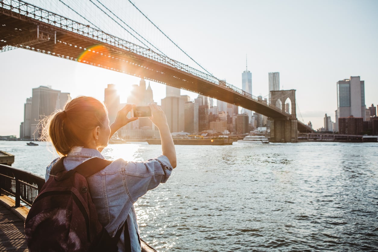A person with a backpack takes a photo of the New York City skyline and Brooklyn Bridge over the East River at sunset. Sunlight shines through the bridge, highlighting the cityscape.