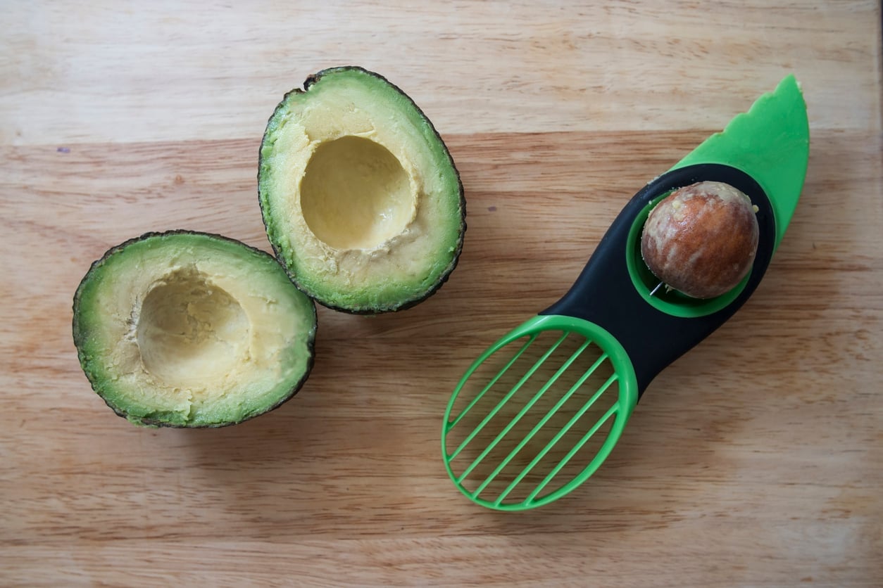 A sliced avocado is on a wooden surface, with one half holding a pit remover tool. This clever kitchen gadget is green and features both an avocado pit extractor and a slotted slicer for cutting the fruit.