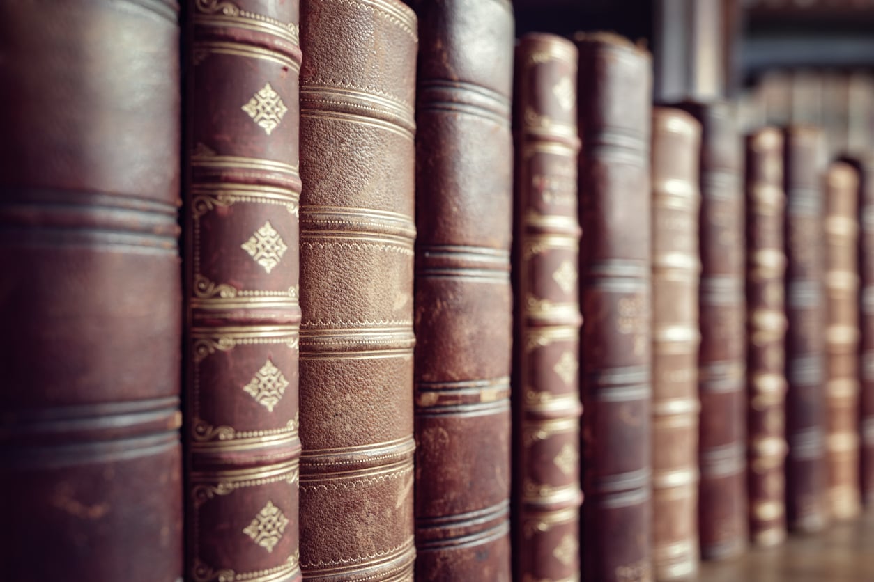 A row of old, leather-bound books with ornate gold detailing on their spines, closely lined up on a bookshelf, evokes the nostalgic charm often found in the living rooms of middle-class moms in the 80s and 90s.