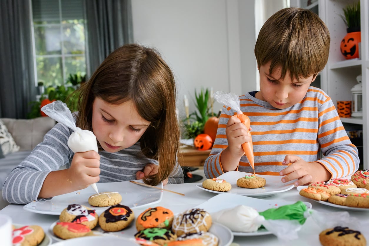 Two children sit at a table decorating cookies with icing, engaging in fun Halloween activities. Halloween-themed cookies are on plates around them, and Halloween decorations are visible in the background.