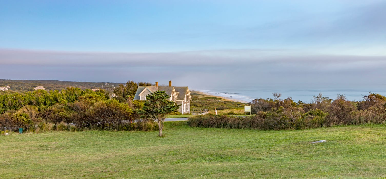 A house sits amid green grass and shrubs overlooking a distant sandy shoreline under a blue sky, with waves visible along the coast—an inviting scene reminiscent of idyllic travel destinations.