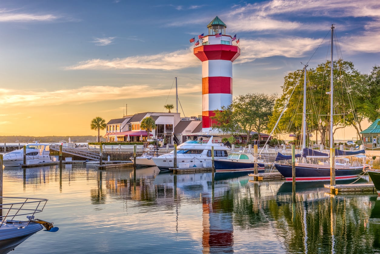 A red and white striped lighthouse stands by a marina with boats docked in calm water at sunset, surrounded by trees and buildings under a partly cloudy sky—a picturesque scene that captures the charm of popular travel destinations.