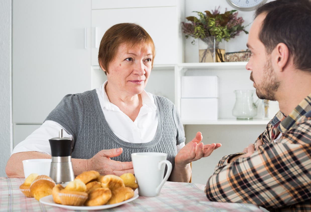 An older woman and a younger man sit at a kitchen table with coffee and pastries, having a serious conversation.