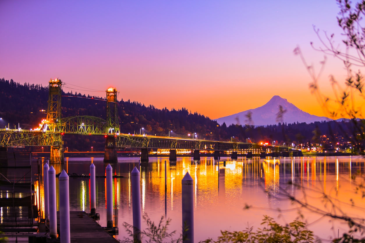 A brightly lit bridge spans a calm river at sunset, with reflections on the water and a snow-capped mountain in the distance. Trees and a colorful sky add to the scenic beauty, making this an unforgettable scene among top travel destinations.