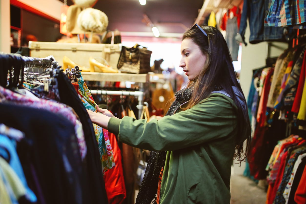 A woman wearing a green jacket browses colorful clothing on racks in a vintage or thrift store, carefully selecting unique pieces rather than waste of money products, with shelves and garments visible in the background.