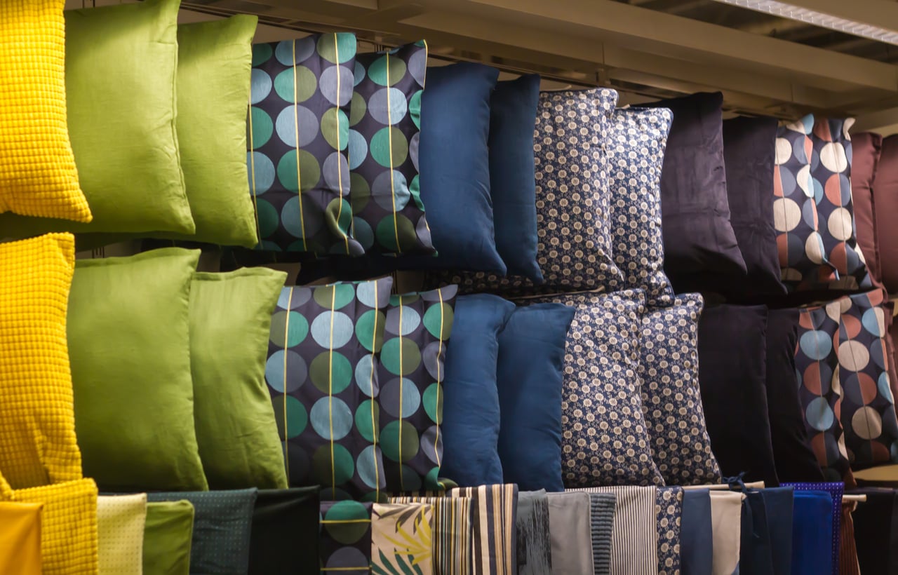 Colorful pillows are displayed on shelves, featuring solid green, blue, and black pillows, as well as others with geometric and patterned designs. Striped and patterned fabric bolts are shown below the pillows.