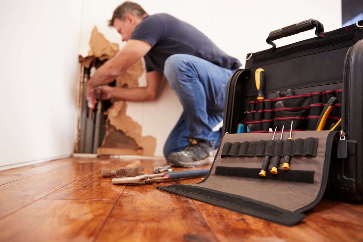 A person repairs pipes inside a wall with exposed plaster, kneeling on a wooden floor. A toolkit with various tools, including screwdrivers and pliers, is open in the foreground—highlighting the benefits of renting with maintenance support.