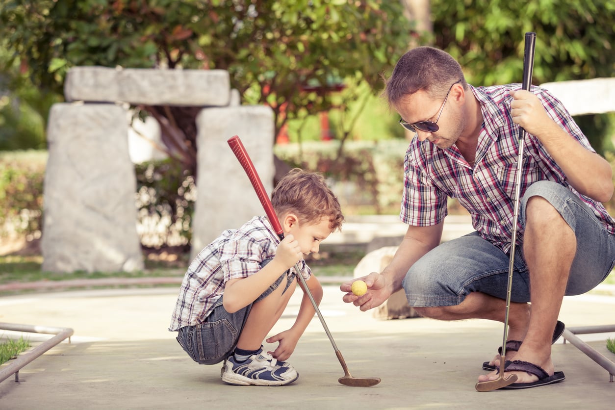 A man kneels beside a young boy, holding a yellow golf ball as they play mini golf. Both wear plaid shirts and shorts. The boy crouches with a putter, focusing on the ball outdoors on a sunny day.