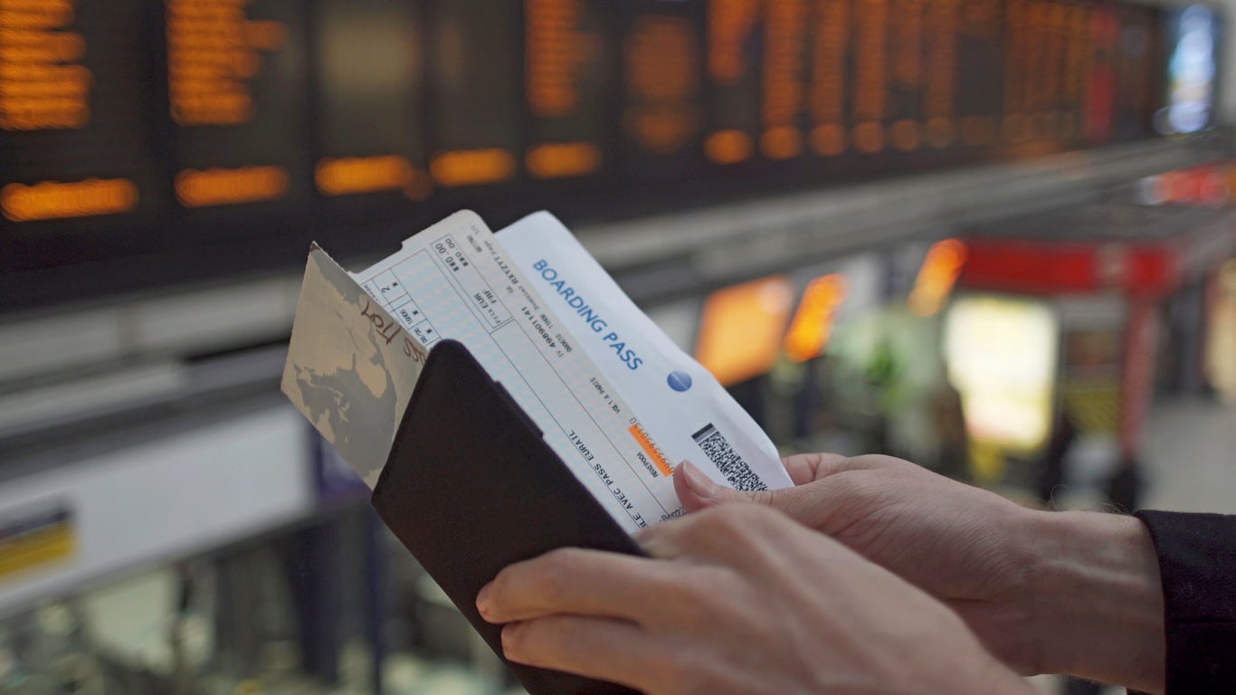 A person holds a passport and boarding pass in an airport, with a large electronic departures board and people blurred in the background, capturing the excitement that builds before booking a flight.