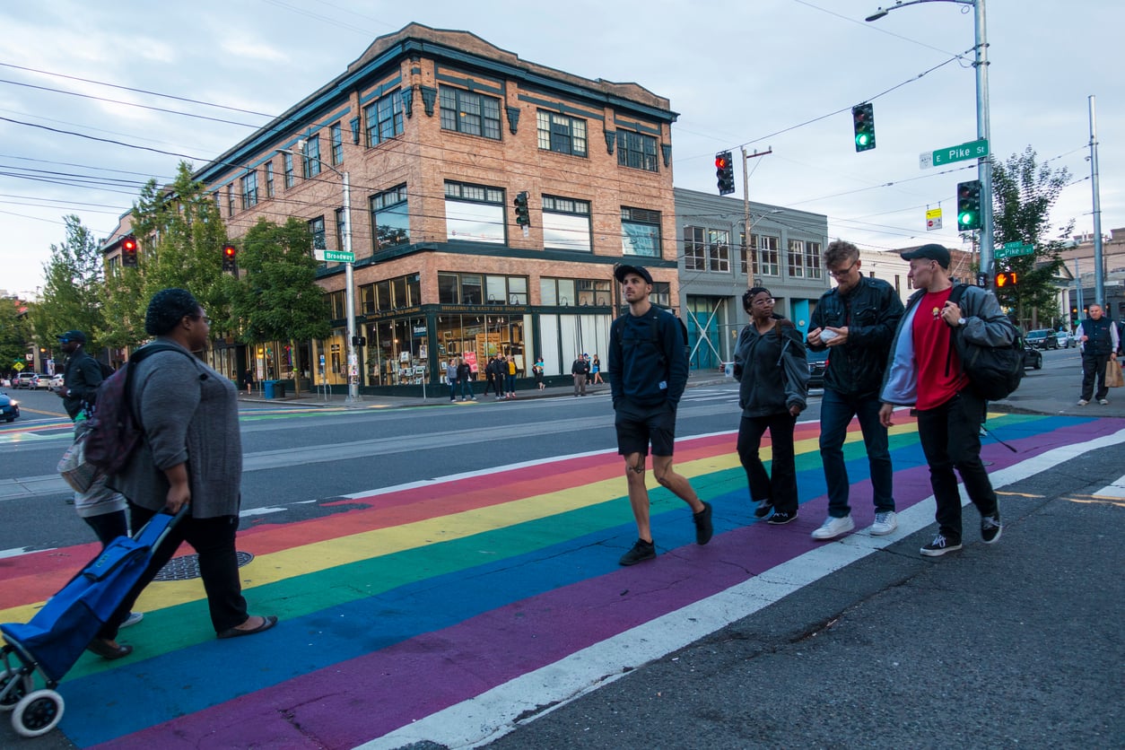 A group of people walk across a rainbow-colored crosswalk at a city intersection with brick buildings and shops in the background. The sky is cloudy and traffic lights are visible.