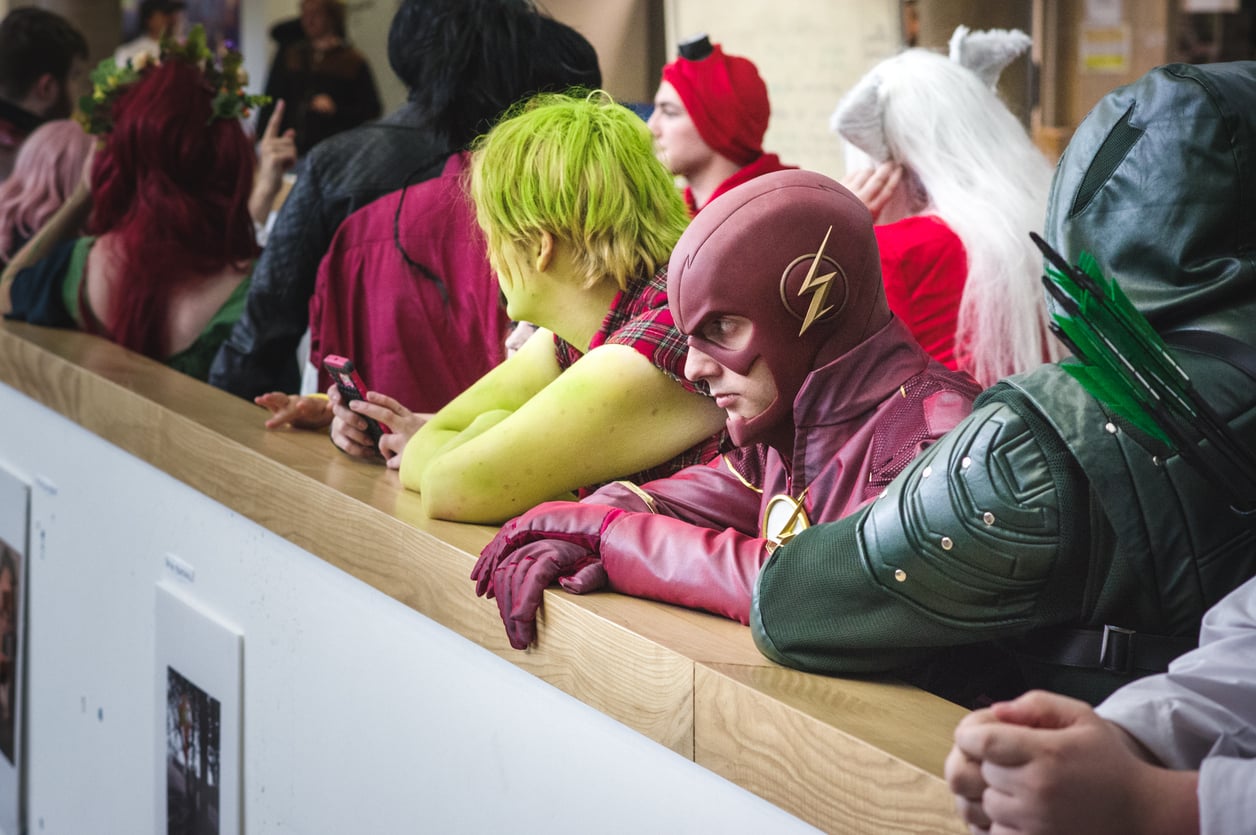 People in superhero costumes, including The Flash and others, lean over a wooden railing at an indoor event. Some are looking ahead while others use their phones. The scene suggests a convention or cosplay gathering, reflecting how expensive hobbies like cosplay bring fans together in vibrant displays of creativity.