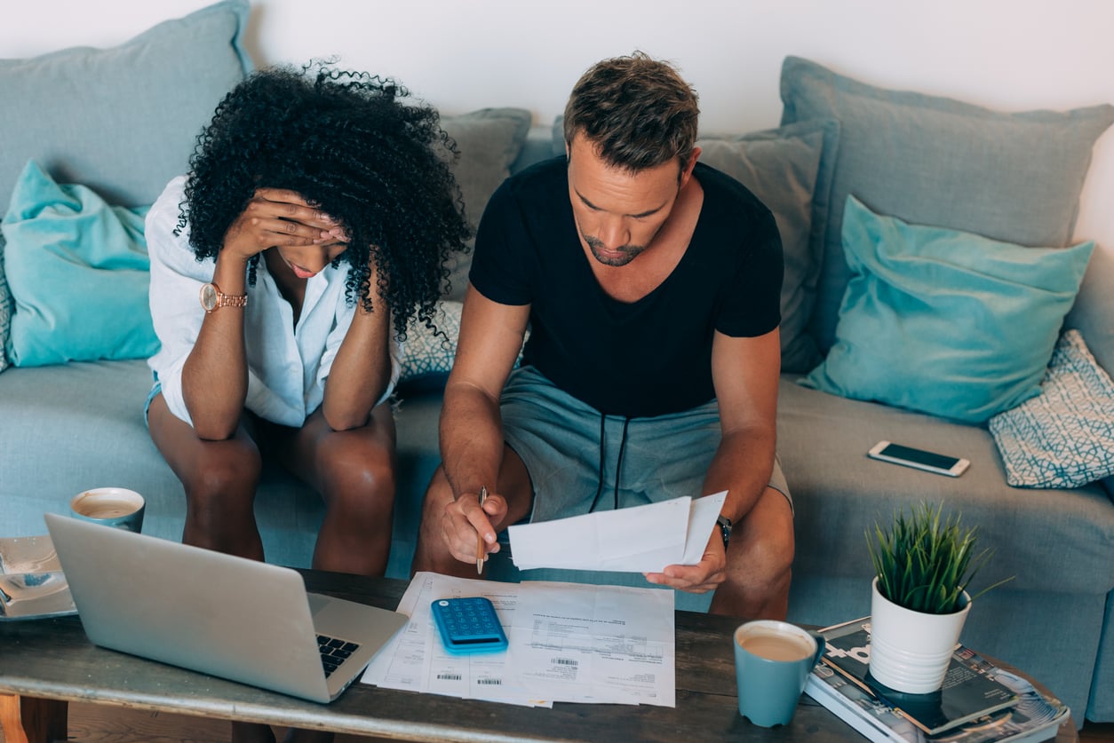 A couple sits on a couch, looking stressed while reviewing bills and paperwork—perhaps reflecting why families in America are having fewer children. A laptop, calculator, and coffee cups sit nearby as they navigate their financial worries together.