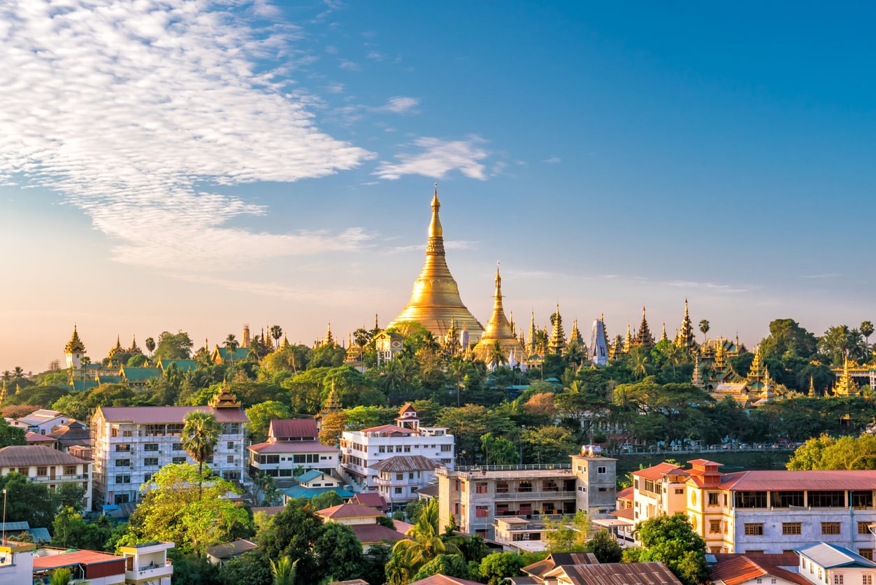 A golden pagoda rises above trees and colorful buildings under a blue sky with scattered clouds, capturing a serene cityscape at sunset—an inviting scene that belongs among the world’s most unforgettable travel destinations.