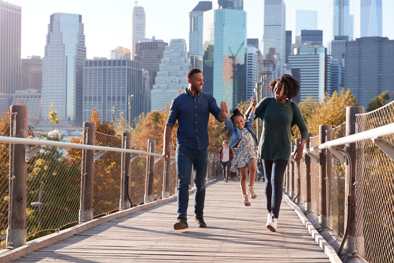 A family of three walks on a bridge in a city during their vacation swap. The parents hold the child's hands and lift her as she smiles, enjoying their time together. Tall skyscrapers and trees are visible in the background on a sunny day.