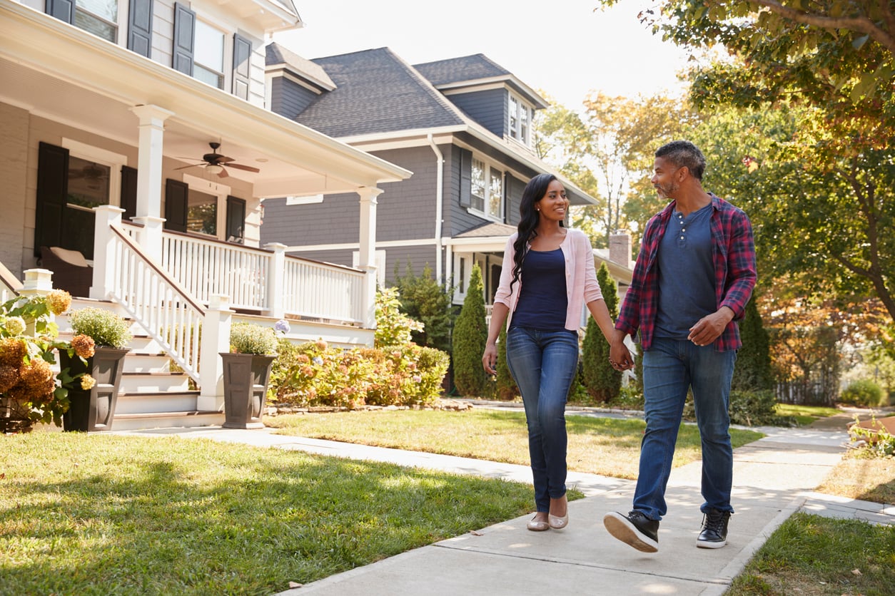 A couple walks hand-in-hand on a residential sidewalk in a suburban neighborhood, smiling and talking about the benefits of renting. Modern homes with porches and manicured lawns are visible in the background on a sunny day.